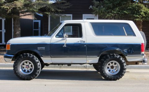 1989 Ford Bronco - A two-tone blue and white Ford Bronco is shown from the side, equipped with large off-road tires.
