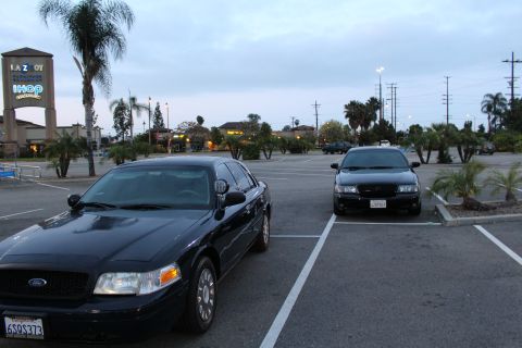 2003 Ford Crown Victoria - dark Ford full-size sedan shown front-on with a Ford grille badge and plate, parked near an IHOP and La-Z-Boy sign at dusk.