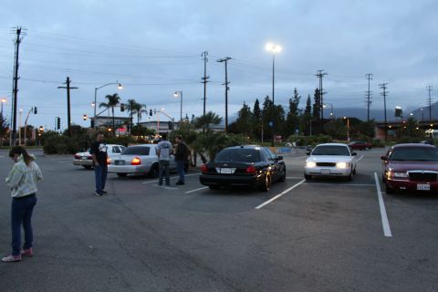 2003 Ford Crown Victoria - few people stand among full-size sedans in an open parking lot at dusk, with headlights on and palm trees in the distance.