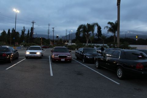 2003 Ford Crown Victoria - row of full-size sedans, including a dark red one, parked in a dimly lit lot at dusk with headlights and palm trees behind.
