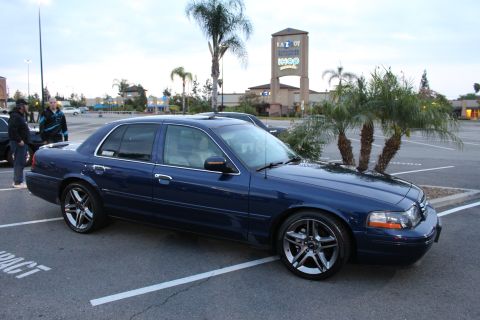 2003 Ford Crown Victoria - dark blue full-size sedan on chrome multi-spoke wheels parked in a lot at dusk, with an IHOP and La-Z-Boy sign behind.