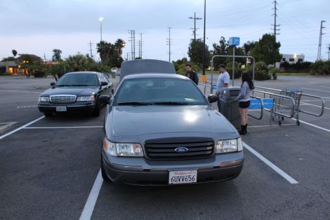 2003 Ford Crown Victoria - gray Ford full-size sedan shown front-on with a Ford grille badge and California plate 6UVV656, parked in a lot at dusk.