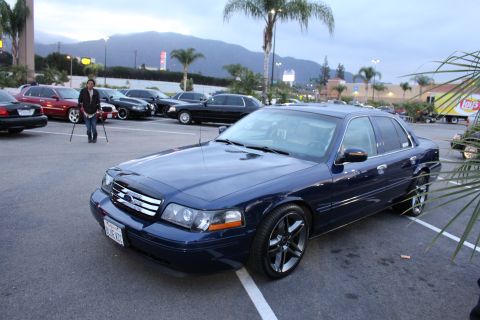 2003 Ford Crown Victoria - dark blue Ford full-size sedan on black alloy wheels with a custom grille, parked in a lot at dusk near mountains and palm trees.
