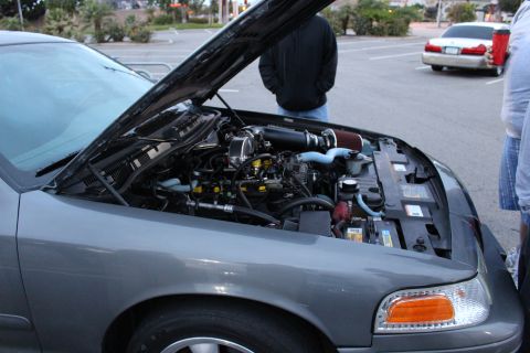 2003 Ford Crown Victoria - open engine bay of a gray full-size sedan showing intake piping and accessories, with people standing behind it at dusk.