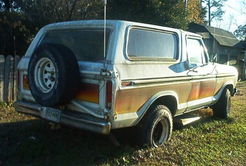 1979 Ford Bronco - Rear three-quarter view of a white late-1970s Ford Bronco parked on grass beside a wood fence and shed, showing the faded orange stripe pac…