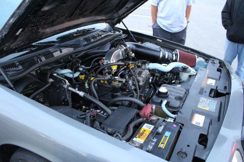 2003 Ford Crown Victoria - Close view of an engine bay with a red cone air filter, intake piping and a battery, under the open hood of a light gray sedan.