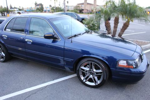 2003 Ford Crown Victoria - dark blue full-size sedan shown in side profile on gray multi-spoke alloy wheels, parked in a lot at dusk by a small palm.