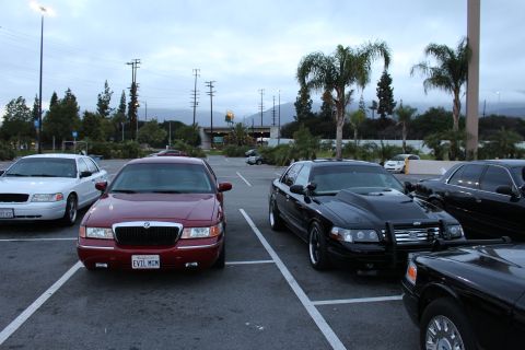 2003 Ford Crown Victoria - dark red Mercury sedan with plate EVIL MGM beside a black Ford sedan with a push bar, parked in a lot at dusk near mountains.