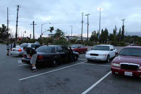 2003 Ford Crown Victoria - Men gather around a black full-size sedan with an open door among parked cars in a lot at dusk, with a dark red Mercury at right.