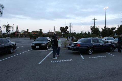 2003 Ford Crown Victoria - Two people stand in marked compact parking spaces between a black sedan and a dark blue Mercury Grand Marquis at dusk.