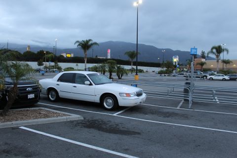 2003 Ford Crown Victoria - white Mercury Grand Marquis sits in a near-empty parking lot beside a black SUV, with mountains and palm trees in the distance.