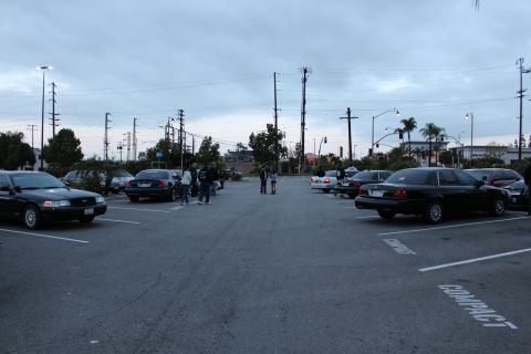 2003 Ford Crown Victoria - wide parking lot at dusk shows black full-size sedans and a small group of people standing among the marked spaces.