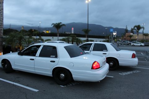 2003 Ford Crown Victoria - Two white Ford Crown Victoria full-size sedans sit parked side by side in a lot at dusk with mountains behind.