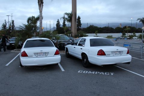 2003 Ford Crown Victoria - Two white Crown Victoria sedans sit parked rear-to-camera in a lot at dusk; one shows a license plate reading GHST VIC.