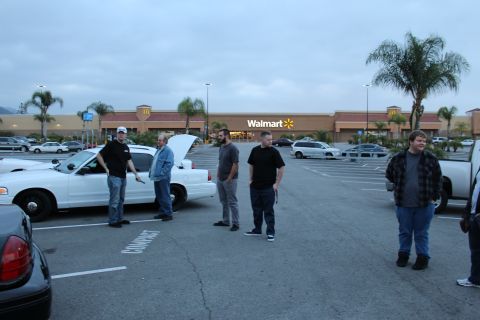 2003 Ford Crown Victoria - People stand around a white sedan with its hood up in a parking lot in front of a Walmart and McDonald's at dusk.