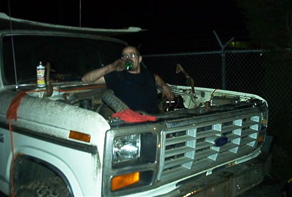 Man sits in the open engine bay of a white 1980s Ford truck with hood removed at night