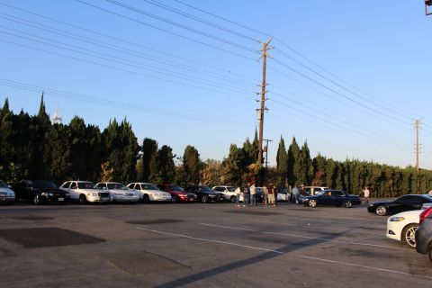 2003 Ford Crown Victoria - Sedans and an SUV park along trees while a few people stand in the wide asphalt lot under a power pole.
