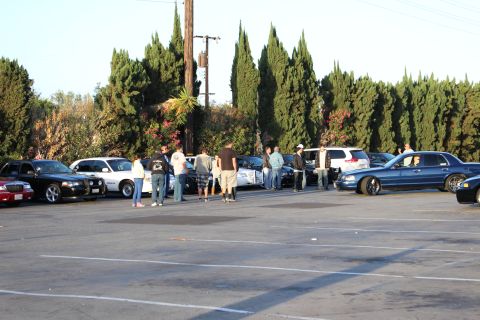 2003 Ford Crown Victoria - cluster of people stand among parked sedans, including a lowered blue sedan at right, beside trees.