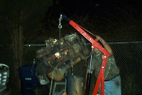 1979 Ford Bronco - An engine is suspended by a red engine hoist, with a man operating it in the background.