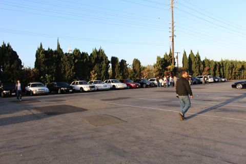 2003 Ford Crown Victoria - man in a jacket walks across the asphalt lot past a long row of parked sedans and gathered people.