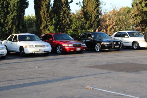 2003 Ford Crown Victoria - white sedan, a red sedan, a black sedan and a tan sedan park side by side facing the camera near trees.