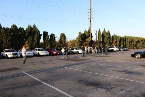 2003 Ford Crown Victoria - People stand and walk in a wide lot beside a row of parked sedans under a clear sky and power pole.