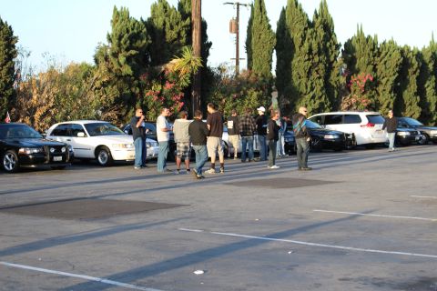 2003 Ford Crown Victoria - group of people stand talking together in a parking lot beside parked sedans and an SUV near trees.
