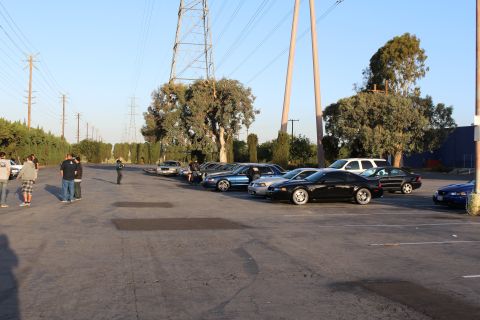 2003 Ford Crown Victoria - line of dark sedans parks diagonally in a wide lot beneath large steel transmission towers and trees.