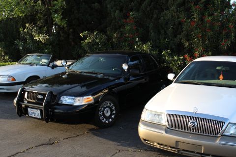 2003 Ford Crown Victoria - black Ford sedan with a chrome push bar and steel wheels parks between a white convertible and a tan Mercury.