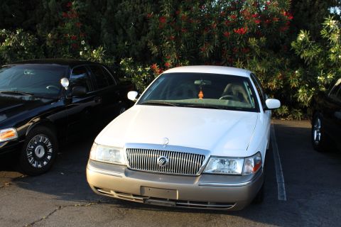 2003 Ford Crown Victoria - white Mercury full-size sedan with a waterfall grille and Mercury emblem parked nose-out next to a black sedan beneath red-flowering bushes.