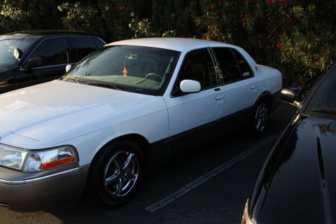 2003 Ford Crown Victoria - Front three-quarter view of a white full-size Mercury sedan with dark lower body cladding and chrome multi-spoke wheels, parked between oth…