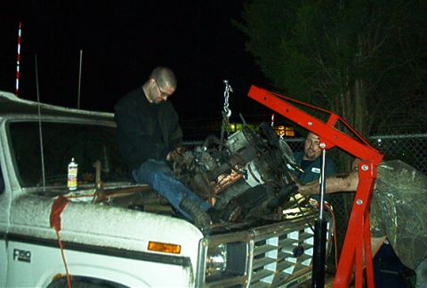 1979 Ford Bronco - Mechanics work on removing an engine from a white truck using a red engine hoist at night.