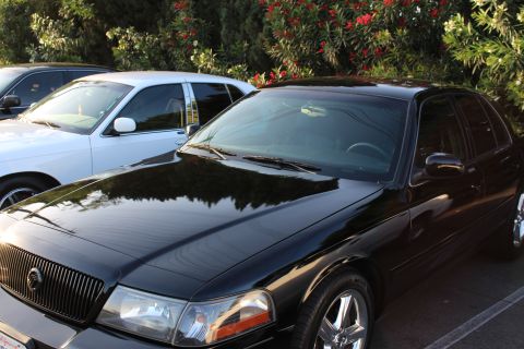 2003 Ford Crown Victoria - Front three-quarter view of a glossy black Mercury sedan with waterfall grille, a white sedan visible behind it beneath a red-flowering bus…