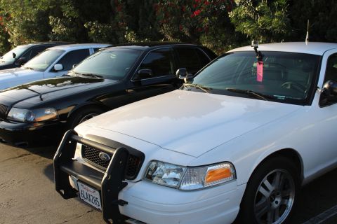 2003 Ford Crown Victoria - white Ford full-size sedan with a black grille guard and California license plate parked nose-out next to two black sedans under shrubs.