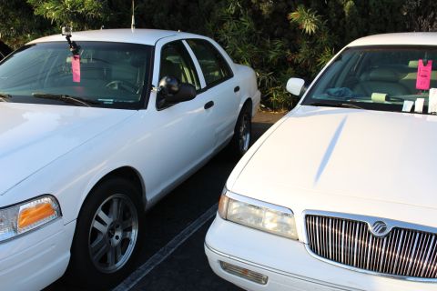 2003 Ford Crown Victoria - Two white full-size sedans parked side by side, the left a Ford and the right a Mercury with a chrome waterfall grille, both with pink wind…
