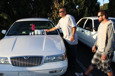 2003 Ford Crown Victoria - Two men stand beside a white Mercury full-size sedan in a lot, one leaning to set papers on the windshield as the other walks past in plaid…