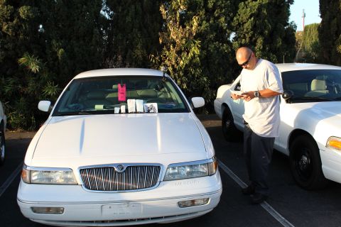 2003 Ford Crown Victoria - man in a white shirt checks his phone beside a white Mercury full-size sedan, with another white sedan parked to the right.