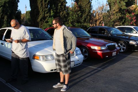 2003 Ford Crown Victoria - Two men stand in a parking lot lined with full-size sedans including white, dark red and black cars, one checking a phone.