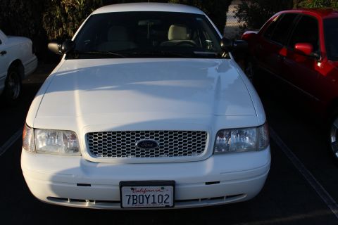 2003 Ford Crown Victoria - Head-on view of a white Ford full-size sedan with a mesh grille, Ford emblem and California plate reading 7BOY102, a red car parked at righ…