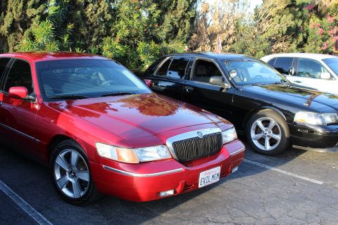 2003 Ford Crown Victoria - dark red Mercury full-size sedan with chrome wheels and a vanity plate reading EVIL MGM, parked beside black sedans under trees.