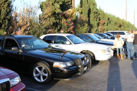 2003 Ford Crown Victoria - row of black and white full-size sedans parked at an angle with a small US flag, several people gathered near the cars at right.