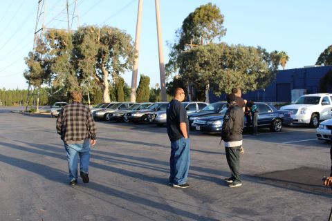 2003 Ford Crown Victoria - Three men stand in an open parking lot looking toward a row of parked dark sedans and a white SUV, with power lines and trees behind.