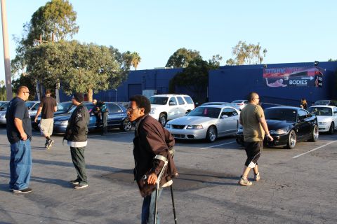 2003 Ford Crown Victoria - Several people, including a man on crutches, walk across a parking lot full of sedans and SUVs in front of a large blue building.