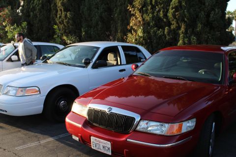 2003 Ford Crown Victoria - dark red Mercury full-size sedan with vanity plate EVIL MGM parked beside a white Ford sedan, a man standing at left in the lot.