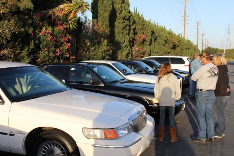 2003 Ford Crown Victoria - group of people stand and talk beside a row of white Mercury Special Edition and dark sedans plus an SUV, lit by low evening sun.