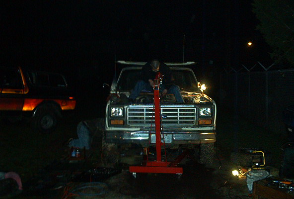 Ford truck with hood open, person working on engine with red lift. Another truck visible to the left
