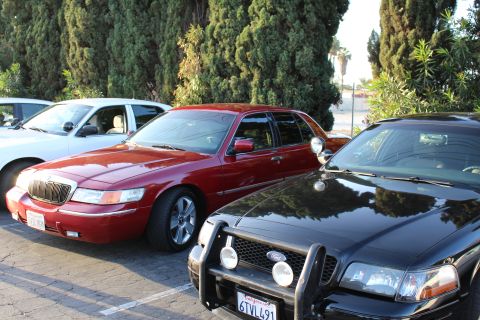 2003 Ford Crown Victoria - dark red Mercury full-size sedan with waterfall grille parked beside a black Ford sedan fitted with a push bar and fog lights, under cypres…