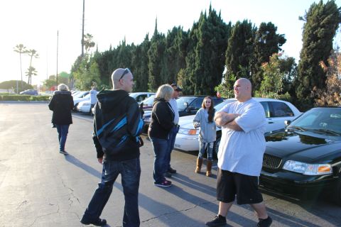 2003 Ford Crown Victoria - group of people stand and chat in a sunlit lot next to parked sedans and tall evergreen trees.