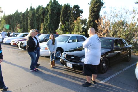 2003 Ford Crown Victoria - People converse in a parking lot beside a black Ford sedan and a white sedan, with evergreens behind.