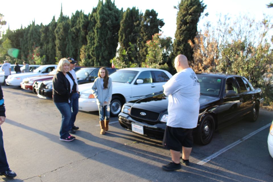 People talking beside a black Ford sedan in a tree-lined lot